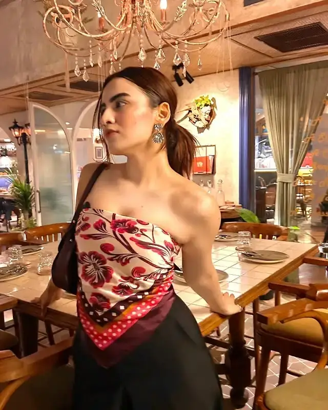 Dubai Call Girls Woman in an off-the-shoulder floral top and red scarf detail poses beside a wooden dining table under a chandelier in a warmly lit restaurant.