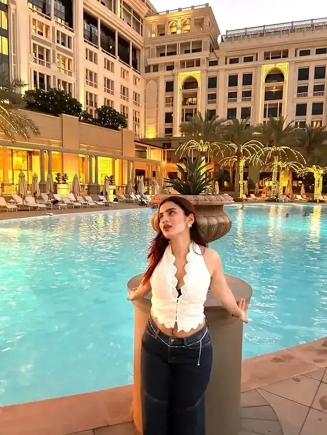 Woman stands by a resort pool with crystal blue water, a grand hotel façade and palm trees in the background, wearing a white halter top and dark jeans.