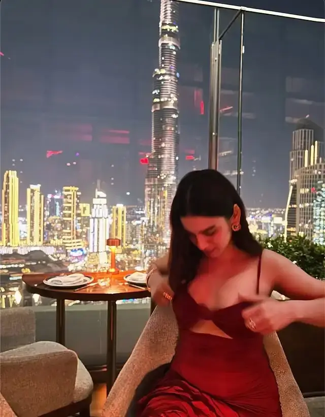 Woman in a red dress at a rooftop restaurant, adjusting her top as a glittering city skyline lights up the night behind her.