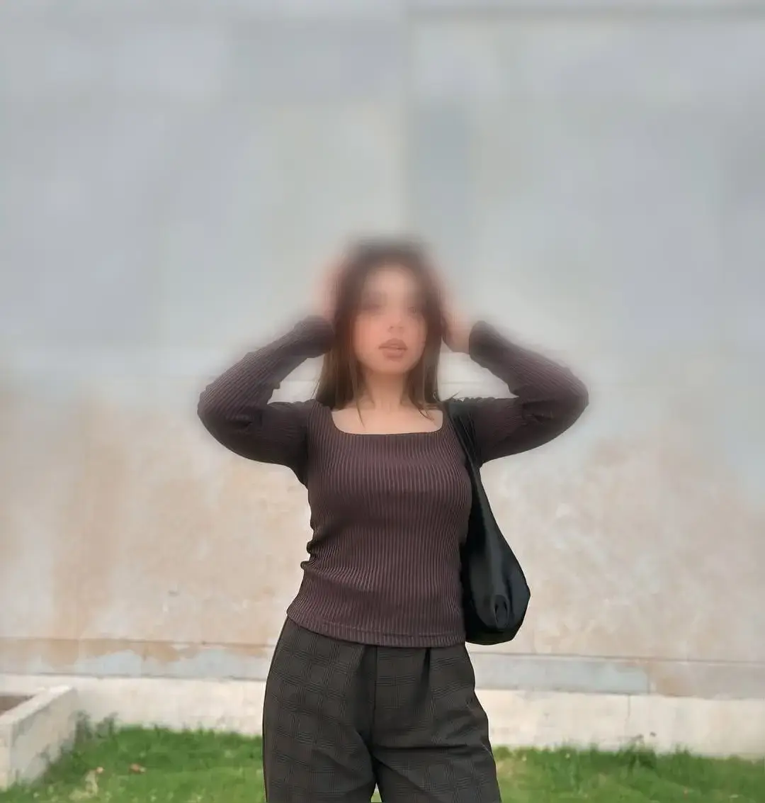 Woman standing outside with hands in her hair, wearing a brown ribbed long-sleeve top and dark plaid pants, carrying a black bag over her shoulder.
